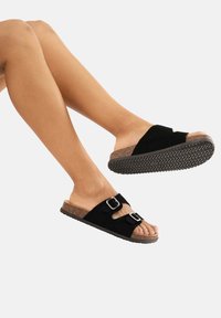 Zwarte suède slip-on sandalen met een kurken voetbed, voorzien van een open teen, dubbele gespaccents en een gestructureerde rubberen loopzool.