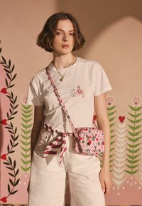 Rosa mønstret crossbody taske med blomstermotiver, justerbar rem og guldfarvede detaljer, båret med en hvid t-shirt og et stribet bælte over hvide bukser.