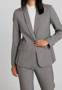 Grau kariertes Blazer mit Reverskragen, zwei Vorderaschen und taillierter Passform. Textilstruktur und minimalistische Designdetails.