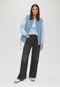 Gepatchte hellblaue Weste über einem weiß-blau gestreiften Hemd, kombiniert mit schwarzen weit geschnittenen Jeans und weißen Sneakers mit braunen Sohlen.
