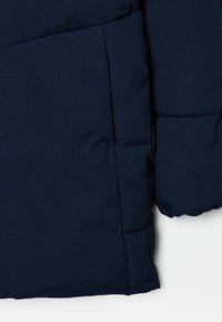 Tmavě modrá vycpaná bunda, na které je vidět boční kapsa a část rukávu s horizontálním prošíváním.