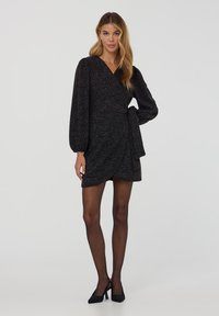 Vestito nero avvolgente con maniche lunghe a sbuffo, tessuto strutturato e cintura in vita. Indossato con calze nere trasparenti e tacchi neri.