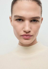 Nahaufnahme eines Porträts mit einem Modell, das eine glatte Haut und natürliches Make-up hat. Es trägt einen hellbeigen hochgeschlossenen Pullover mit weicher Textur.