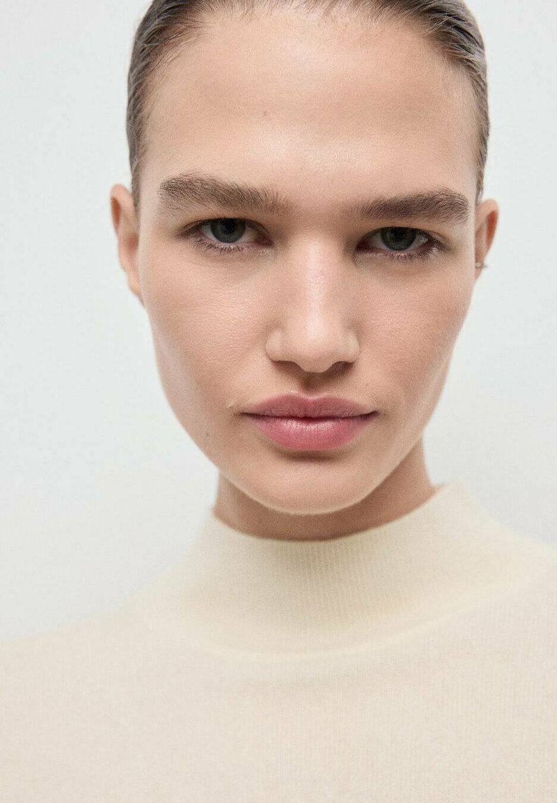 Nahaufnahme eines Porträts mit einem Modell, das eine glatte Haut und natürliches Make-up hat. Es trägt einen hellbeigen hochgeschlossenen Pullover mit weicher Textur.