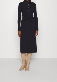 Marinblå långärmad klänning med krage och knäppning fram. Inkluderar ett midjeband och är tillsammans med ljusbeige knähöga stövlar.