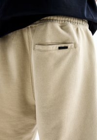 Pantalon de survêtement beige avec taille élastique et une poche arrière arborant une petite étiquette noire "PULL&BEAR", porté avec un top noir.