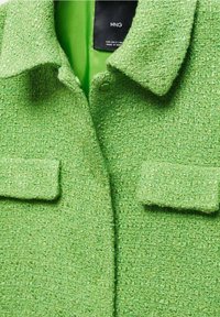 Giacca verde tessurizzata con un motivo intrecciato, dotata di due tasche frontali e un colletto, foderata in tessuto verde uniforme.