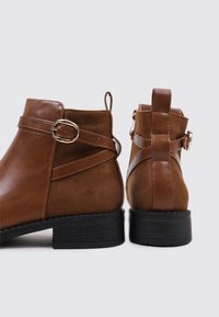 Botas de tobillo de cuero marrón con un acabado suave, que cuentan con una correa decorativa y un accentuador de hebilla, y suelas de goma negra.