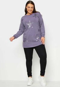 Lila ribbad tröja med paljetter i stjärnmönster, långa ärmar och en avslappnad passform, kombinerad med svarta tajta jeans och vita sneakers.