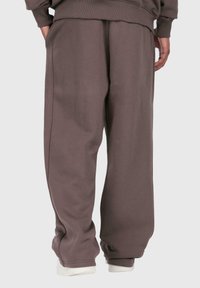 Person in lockeren braunen Sweatpants mit den Händen in den Taschen, der vor einem schlichten hellen Hintergrund steht.
