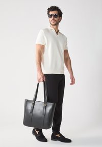 Black leather tote bag with two sturdy handles, smooth texture, and gold hardware accents. Model wears a light beige polo and black pants.