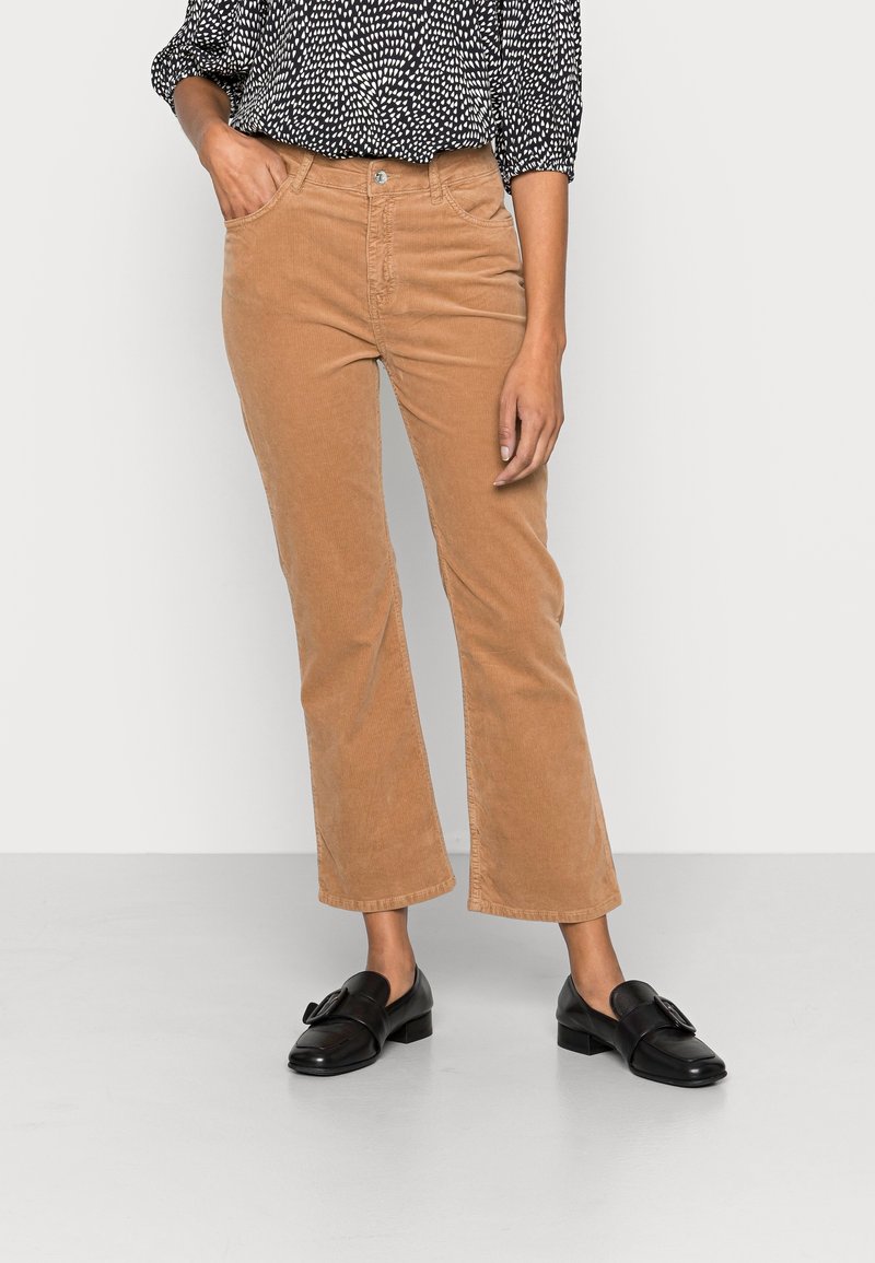 Femme portant un pantalon en velours côtelé beige, des mocassins noirs avec des nœuds et un chemisier à manches longues noir et blanc à motifs, sur un fond uni.