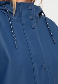 Αδιάβροχο μπουφάν βροχής σε ναυτικό μπλε χρώμα με ομαλή υφή, κουμπιά τύπου κλίπ και κουκούλα με κορδόνι που διαθέτει αντίθετες λευκές λεπτομέρειες.