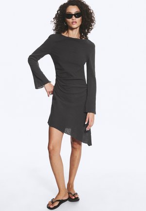 Femme aux cheveux bouclés portant une robe asymétrique noire, des lunettes de soleil noires et des sandales noires, debout avec une main sur la hanche.