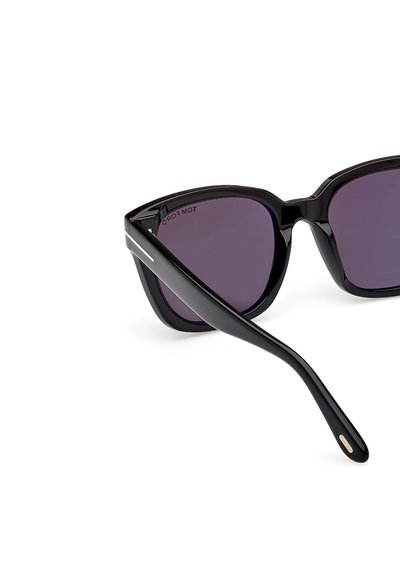 Lunettes de soleil à monture noire avec une forme angulaire et des verres violets foncés. Elles présentent une finition brillante et des accents en métal argenté sur les branches.