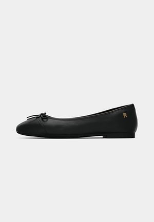 Musta nahkainen ballerina-malli, jossa on pieni rusetti edessä ja kultainen logo kantapään lähellä, esitetty sivuprofiilissa valkoista taustaa vasten.