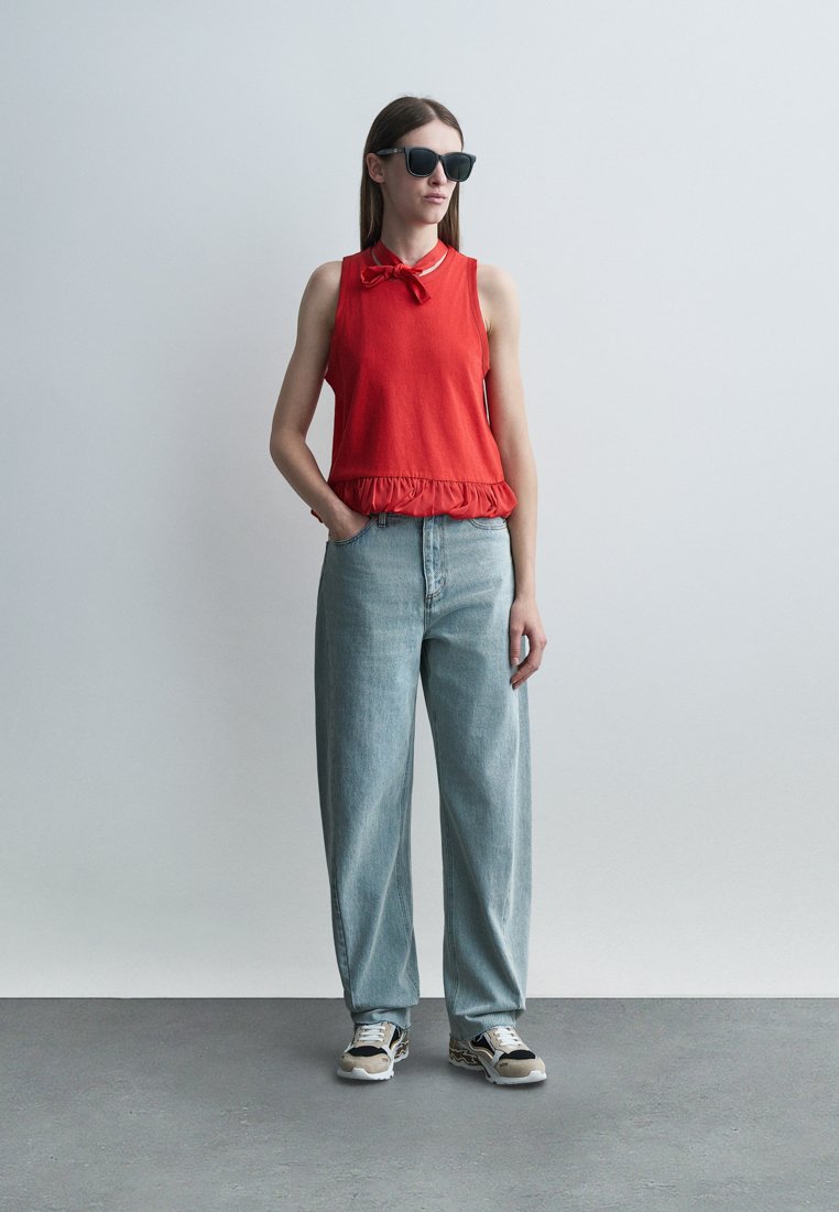 Femme portant des lunettes de soleil noires, un top sans manches rouge avec un nœud, un jean bleu clair ample et des baskets, debout avec une main dans la poche.
