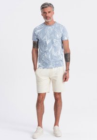 Camisa de manga corta azul claro con patrones de hojas blancas, combinada con pantalones cortos crema y zapatillas blancas, exhibida en un fondo neutro.