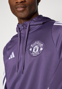 Violetti urheiluhuppari, jossa on vetoketju ja nyörit. Hankkeessa valkoiset Adidas-raidat ja Manchester Unitedin logo rinnassa. Sileä kangasrakennus.