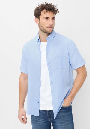 Mann trägt hellblaues kurzärmeliges Hemd über weißem T-Shirt mit blauer Jeans und steht mit einer Hand in der Tasche.