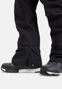 Zwarte waterdichte broek met ritsmanchetten, gecombineerd met stevige zwarte laarzen met een gestructureerde zool en witte accenten. Vibram-logo zichtbaar.