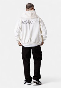 Weißer Hoodie mit dem aufgenähten Schriftzug "estéem" auf dem Rücken, lässige Passform, lange Ärmel und Kapuze mit Kordelzug. Darunter werden schwarze Cargo-Hosen getragen.