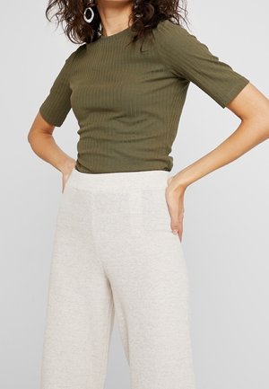 Mujer con camiseta de manga corta acanalada de color verde oliva y pantalones anchos de punto crema de talle alto, con las manos apoyadas en las caderas.