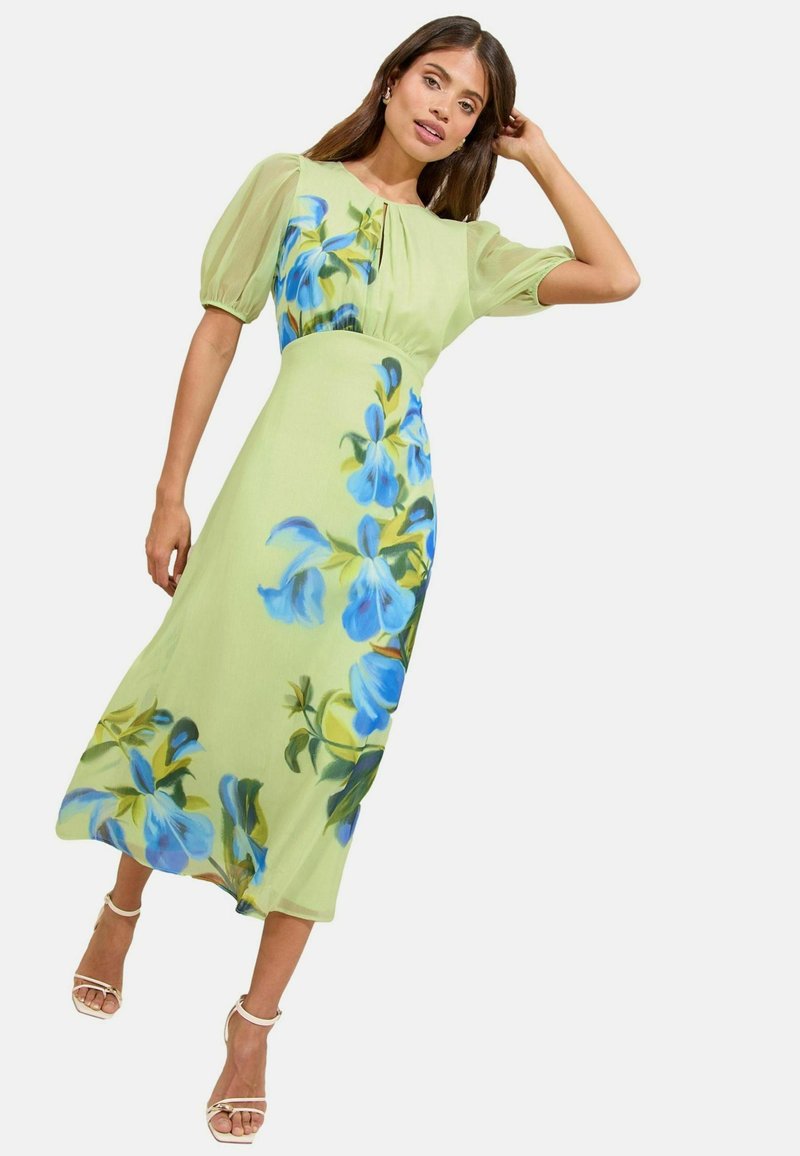 Groene bloemen midi-jurk met een aansluitende lijfje, pofmouwen, blauwe bloemenpatronen en een zwierige rok. Gecombineerd met bandjeshakken.