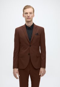 Bruin pak met een slim fit, zwart shirt eronder, enkele knoopsluiting en een borstzak met een donkere driehoekige accent. I