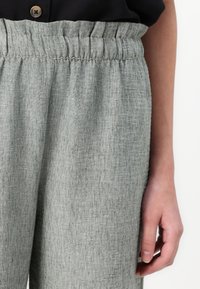 Světlé zelené kalhoty z texturované látky s elastickým pasem. Detailní pohled zaměřený na oblast pasu a umístění ruky.