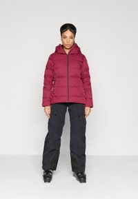 Casaco puffer bordo com capuz e fecho na frente, combinado com calças de carga pretas. O casaco apresenta um design acolchoado e um logotipo no peito.