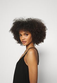 Abito nero con spalline sottili, realizzato in un tessuto morbido e fluido. Il soggetto ha i capelli ricci e indossa una delicata collana a catena.