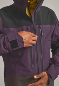 Purple jacket with black accents, zippered pockets, and a hood. Features a velcro cuff and a yellow lining for insulation.