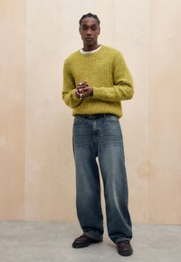 Suéter amarelo tricotado, calças de ganga azuis oversized e calçado escuro. Mãos entrelaçadas, fundo simples com painéis de madeira clara.