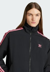 Zwarte windjack met hoge kraag, roze zijstrepen en geborduurd logo. Heeft een ritsluiting en een gladde textuur.