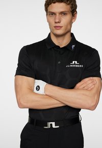 Black polo shirt with hexagonal pattern, featuring a logo on the chest and collar. Worn with a black belt, and white golf glove on the left hand.