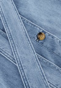 Lichtblau denim stof met diagonale naden en een bruine knoopdetail. De textuur toont de typische denimweving en stiksels.