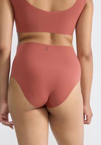 Kvinna som bär sömlösa högmidjade underkläder i rostfärg och matchande bh, visad bakifrån mot en enkel bakgrund.