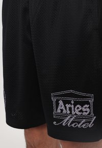 Shorts en maille noire avec un logo graphique blanc "Aries Motel" bien en vue. Le tissu a une texture respirante et une coupe ample.
