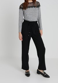 Maglietta a maniche lunghe grigia con sovrapposizione in rete nera e rifiniture in pizzo, abbinata a pantaloni neri con vita elastica. Il modello indossa mocassini neri con dettaglio a catena.