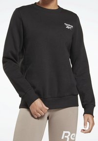Sudadera negra con cuello redondo y puños acanalados. Presenta un logo blanco de Reebok en el pecho izquierdo. Tejido suave y texturizado.
