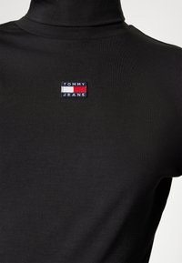 Svart polo som är gjord av slät tyg, med en liten broderad "TOMMY JEANS"-logotyp med röda och vita detaljer.