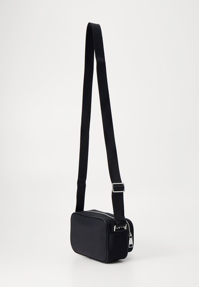 Sac bandoulière noir de forme rectangulaire, fabriqué en tissu doux, avec une fermeture éclair et une sangle réglable, rehaussé de matériel en argent.