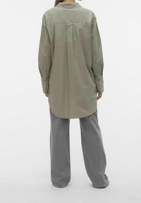 Olijfgroene blouse met een achterpaneel en lange mouwen, gecombineerd met losse grijze broek in een zachte, gestructureerde stof.