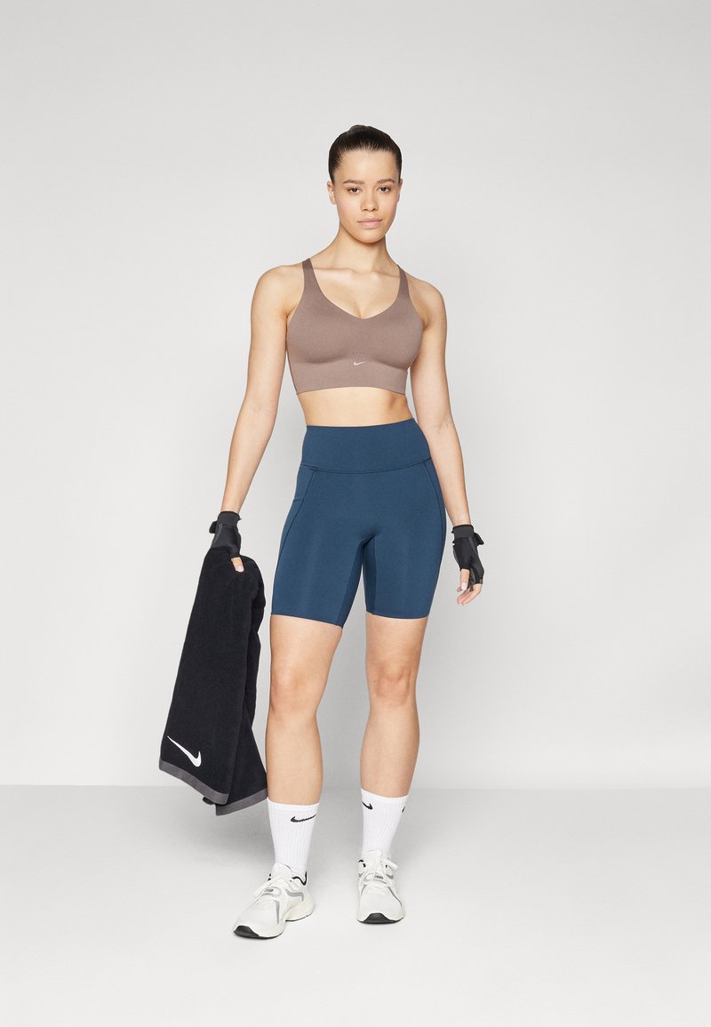 Braun Sport-BH, marineblaue Shorts, schwarzes Handtuch und weiße Socken. Glatte Textur, figurbetonter Stil mit sichtbaren Markenlogos. Sportliches Schuhwerk.