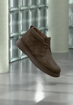 Καφέ καστόρινη μποτάκια αστραγάλου με κορδόνια που αιωρείται πάνω από ένα ανακλαστικό πάτωμα με φόντο ένα παγωμένο γυάλινο παράθυρο και κολόνα.