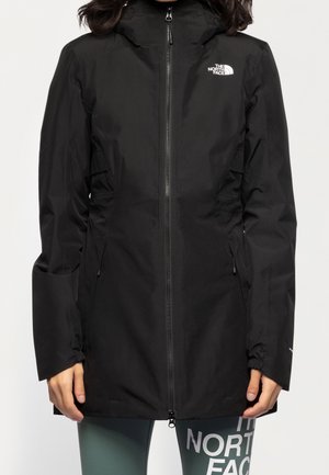 Femme portant une veste imperméable noire North Face avec une fermeture éclair frontale et une capuche, associée à un legging vert portant le texte blanc North Face.
