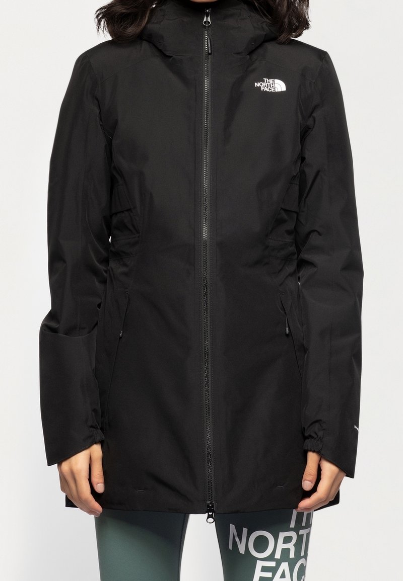 Femme portant une veste imperméable noire North Face avec une fermeture éclair frontale et une capuche, associée à un legging vert portant le texte blanc North Face.