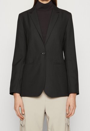 Femme portant un blazer noir cintré à un bouton sur un col roulé noir, associé à un pantalon beige clair avec des poches latérales.