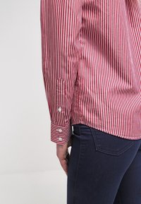 Personne portant une chemise rayée rouge et blanche à manches longues et un jean bleu foncé, vue de côté au niveau de la taille et du bras.